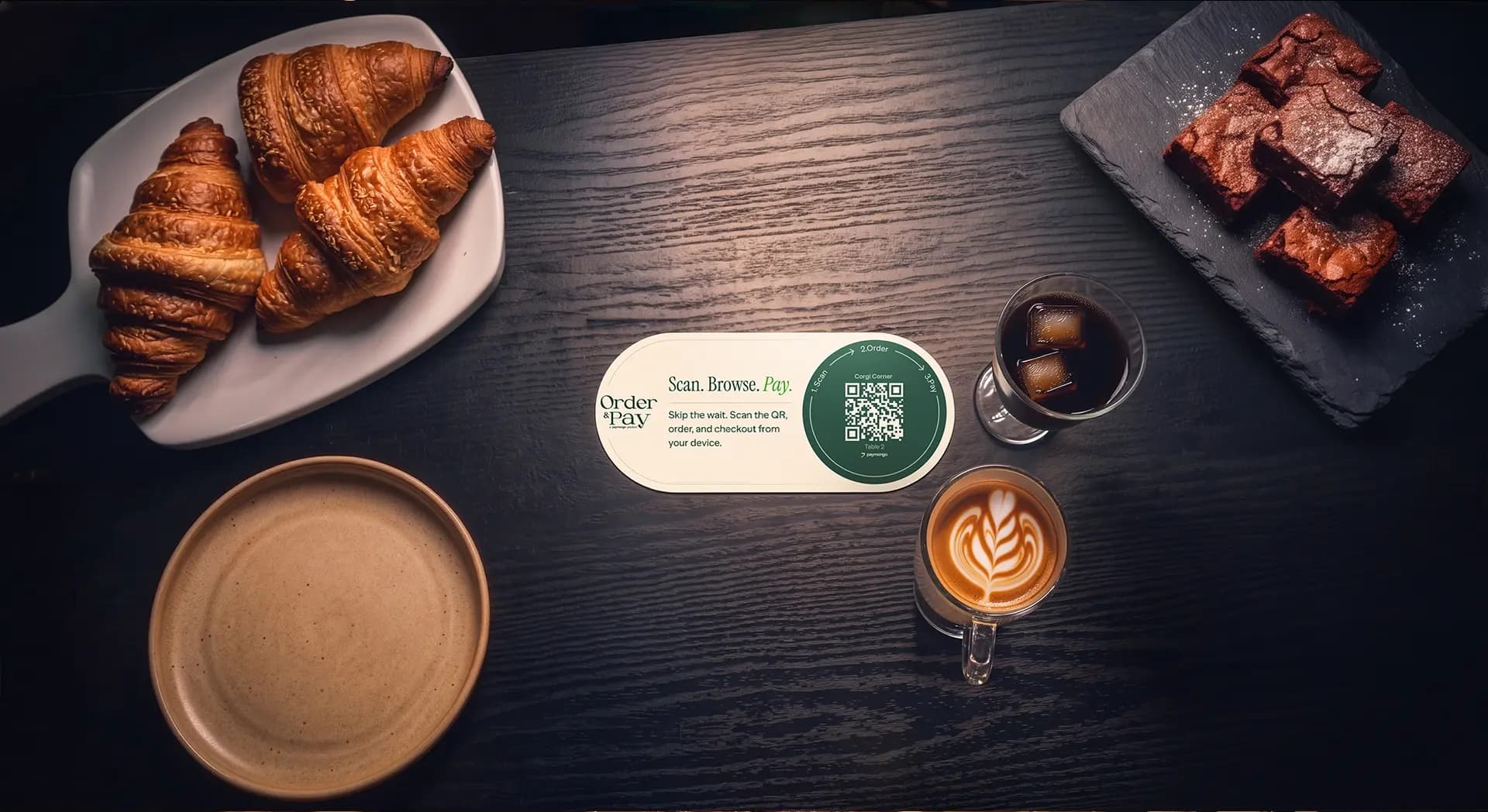 An Order and Pay QR code card on a dark wood café table, beside croissants, brownies, an iced coffee and a latte — customers scan it to open the menu, order, and pay from their own phone