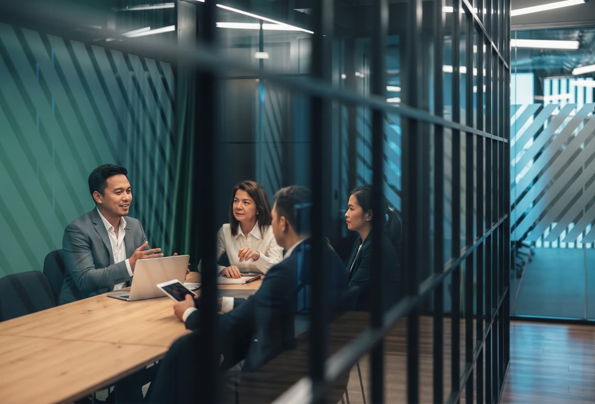 Four colleagues in business attire meet around a table in a glass-walled conference room