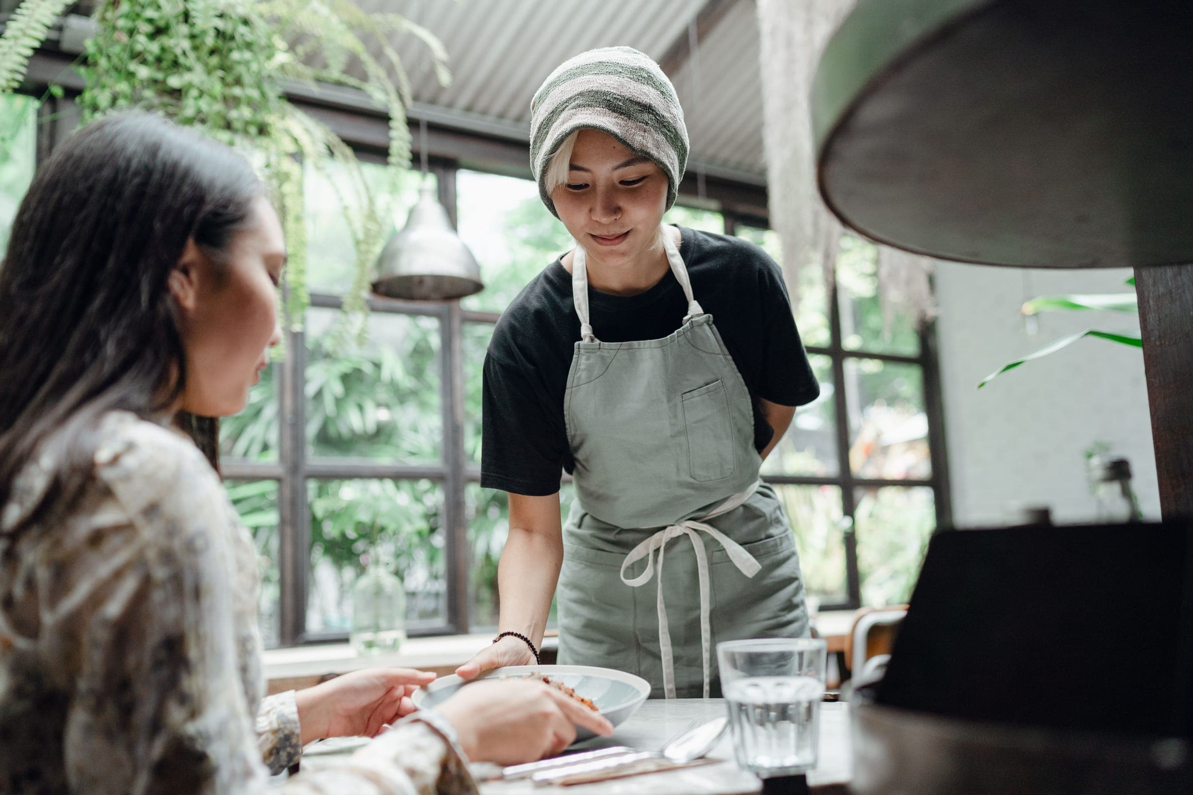 A smiling server in an apron sets a bowl down in front of a guest at a café table