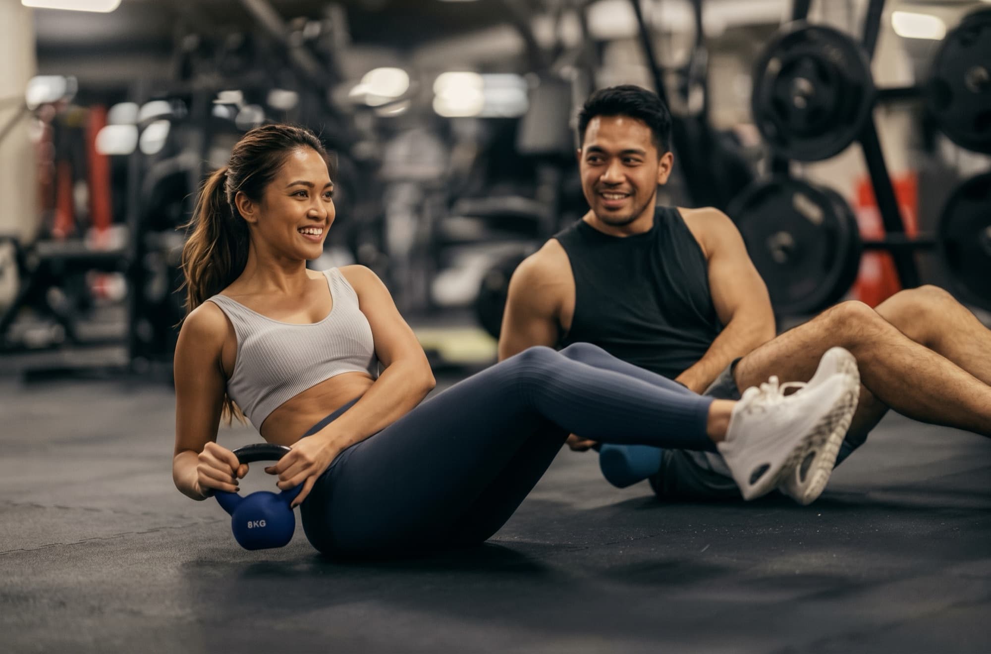 A woman holds a kettlebell in a seated twist beside a smiling training partner on a gym floor